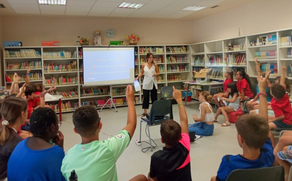 Fotografía del taller de la biblioteca de Cintruénigo, tomada desde el final de la sala. En la que aparecen los menores asistentes de espaldas levantando la mano y la chica que está dando la formación sonriendo. 