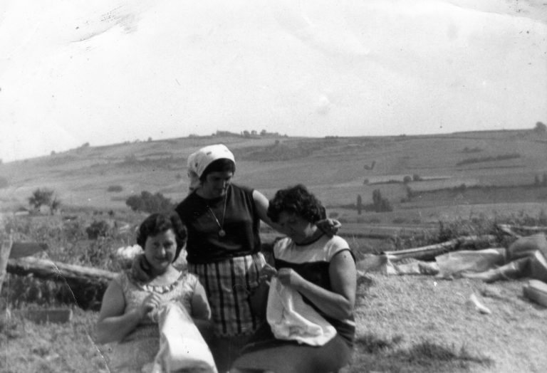 Fotografía en blanco y negro en la que aparecen tres mujeres cosiendo ropa en un campo. 