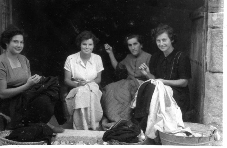 Fotografía en blanco y negro en la que aparecen cuatro mujeres cosiendo ropa en la puerta de una casa. 