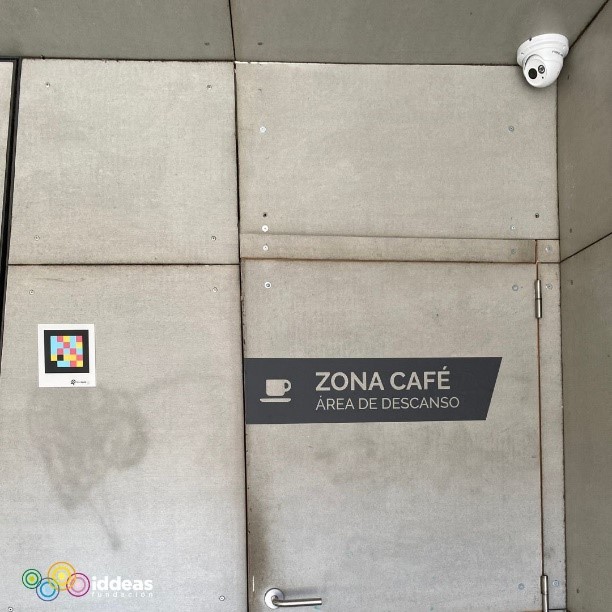 Puerta de acceso a la cocina, de hormigón gris, cerrada y señalizada con un cartel que pone: Zona de café. Área de descanso y el símbolo de una taza. A su izquierda un código NaviLens y arriba a la izquierda una cámara de seguridad.