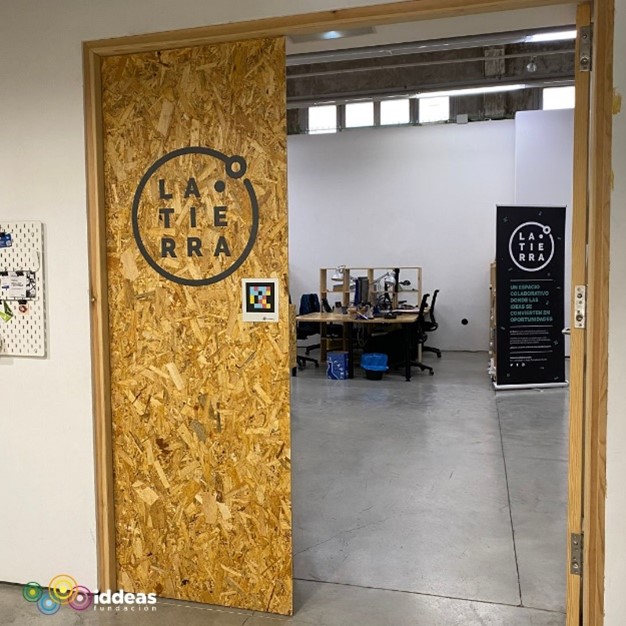 Puerta de acceso al espacio de trabajo abierta a una parte del interior del coworking, donde se ven unas mesas y unas sillas. En la puerta podemos observar el logo de 