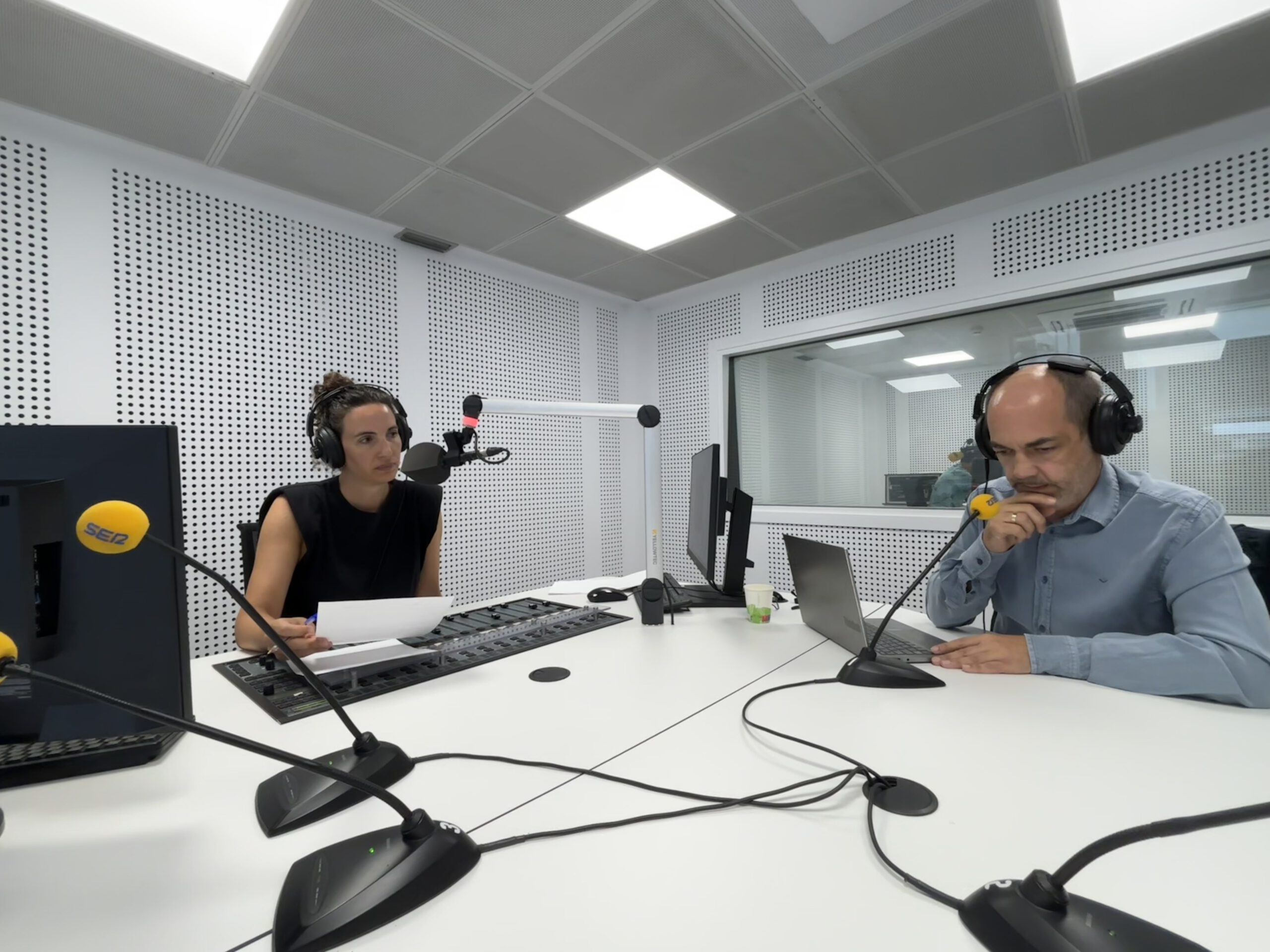 Dos personas con auriculares y micrófonos de la cadena SER en una cabina de radio insonorizada. Están sentadas frente a una mesa con consolas de sonido, papeles y un ordenador.