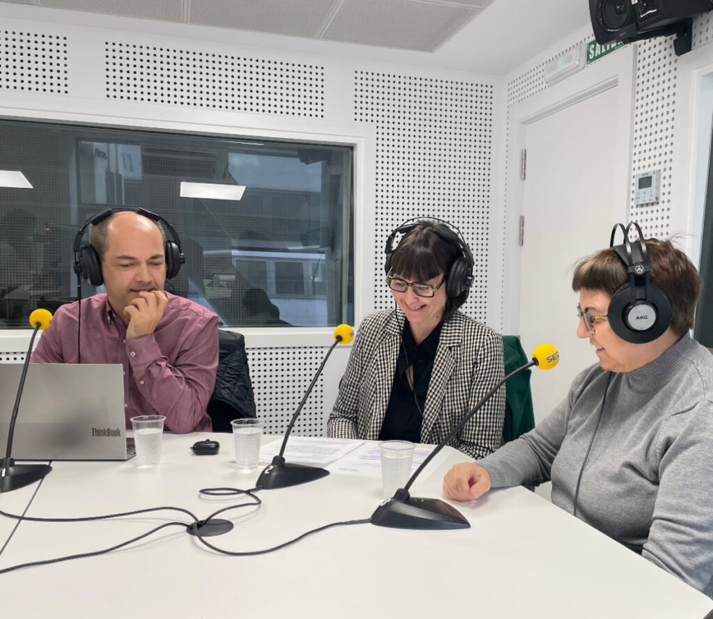 Tres personas con auriculares y micrófonos de la cadena SER en una cabina de radio. Están sentadas alrededor de una mesa blanca con documentos, vasos de agua y un ordenador portátil.