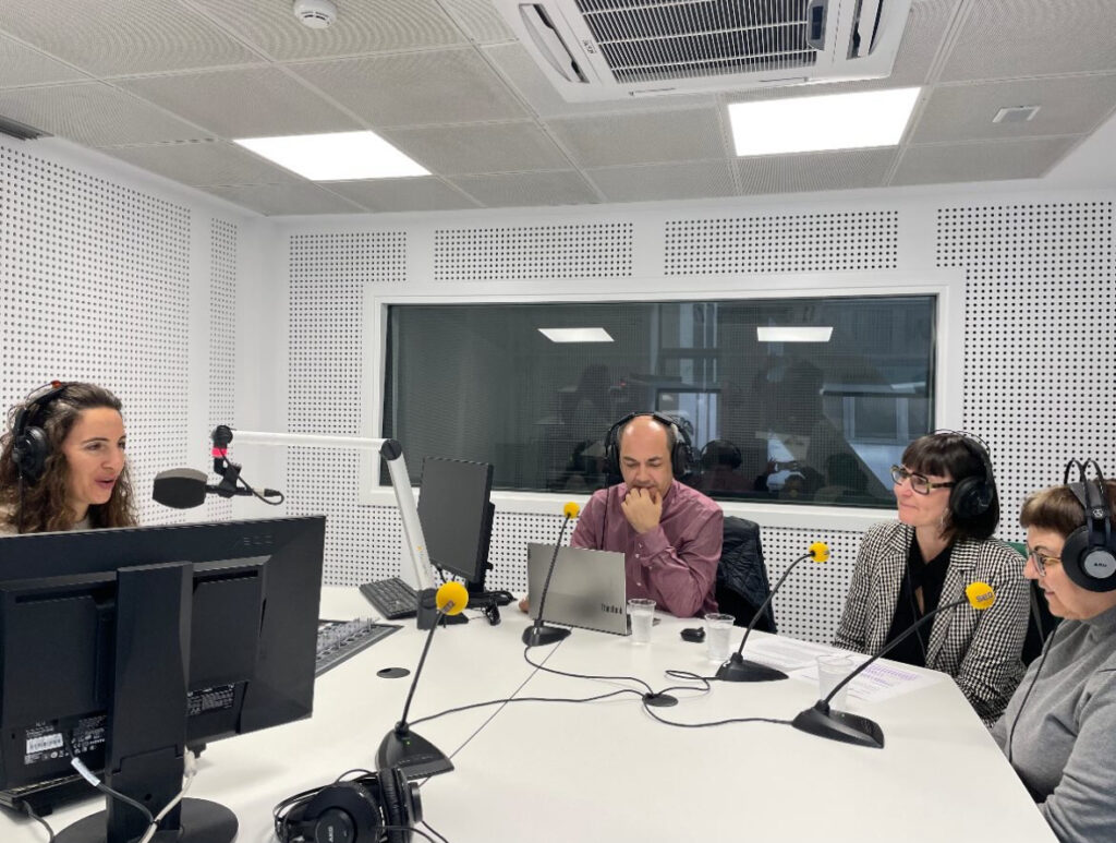 Cuatro personas con auriculares y micrófonos en una cabina de radio de la cadena SER. Tres están sentadas frente a una mesa con documentos, ordenadores y vasos de agua, mientras una cuarta persona, situada frente a ellas, conduce la conversación desde una posición con doble monitor y micrófono.
