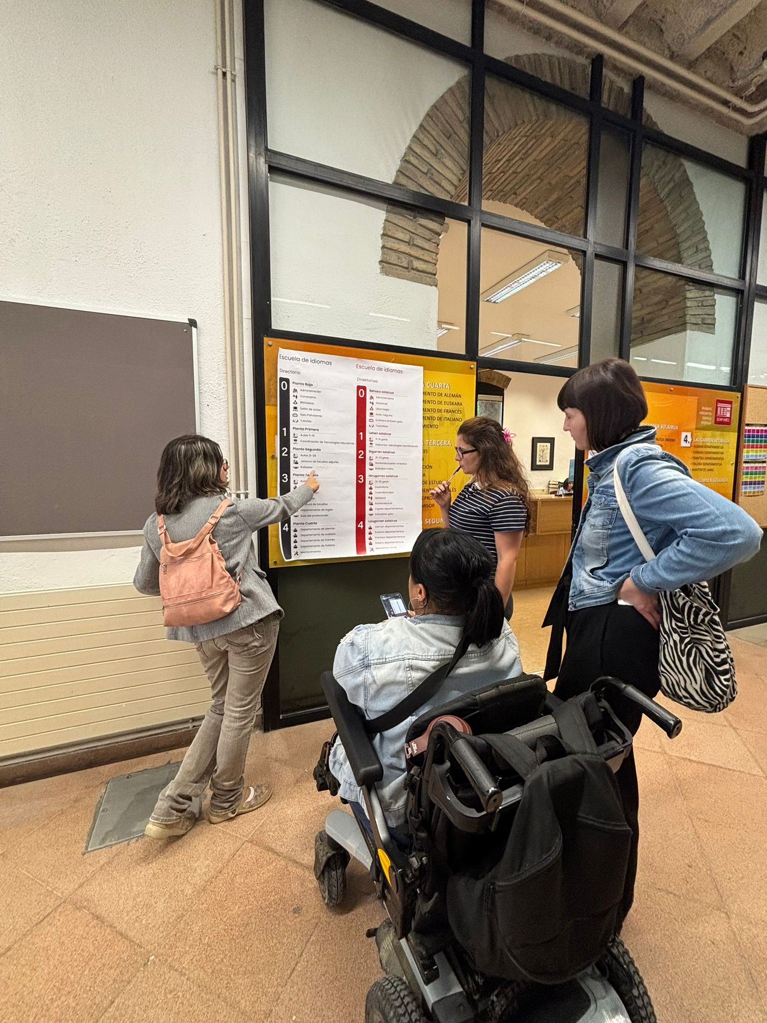 Cuatro personas observan un directorio informativo en la entrada de un edificio. Una de ellas señala la información del cartel, mientras otra utiliza una silla de ruedas eléctrica. El grupo conversa frente a la señalización, que indica las plantas y servicios disponibles en la escuela de idiomas.