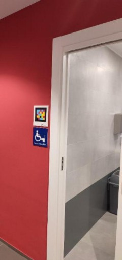 Pared roja junto a una puerta blanca con cristal traslúcido; al lado del marco, dos señales pequeñas: arriba un código NaviLens de colores y debajo un pictograma de accesibilidad (silla de ruedas) en azul. Al fondo se intuye un espacio interior en tonos grises.