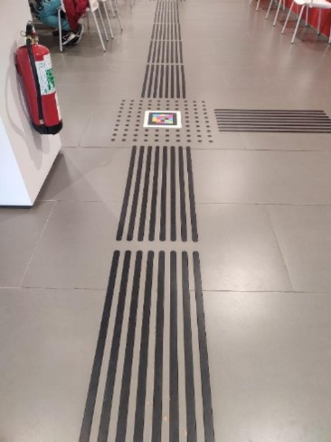 Suelo de baldosas grises en un interior, con un encaminamiento podotáctil negro: bandas longitudinales y una zona de botones en relieve que marcan un cruce. En el centro del cruce hay un código NaviLens cuadrado de colores. A la izquierda se ve un extintor y, al fondo, mesas y sillas.