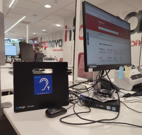 Mostrador de atención en una oficina abierta: sobre la mesa hay un ordenador con monitor encendido, teclado y cables. En primer plano se ve un equipo negro con el pictograma de bucle magnético (accesibilidad auditiva) y al fondo aparecen puestos de trabajo.