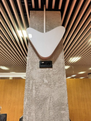 Columna de piedra en un interior, con un dispositivo Visualfy en forma de corazón blanco instalado en la parte alta y, debajo, un pequeño panel negro. Al fondo se ve un techo con lamas de madera e iluminación integrada.