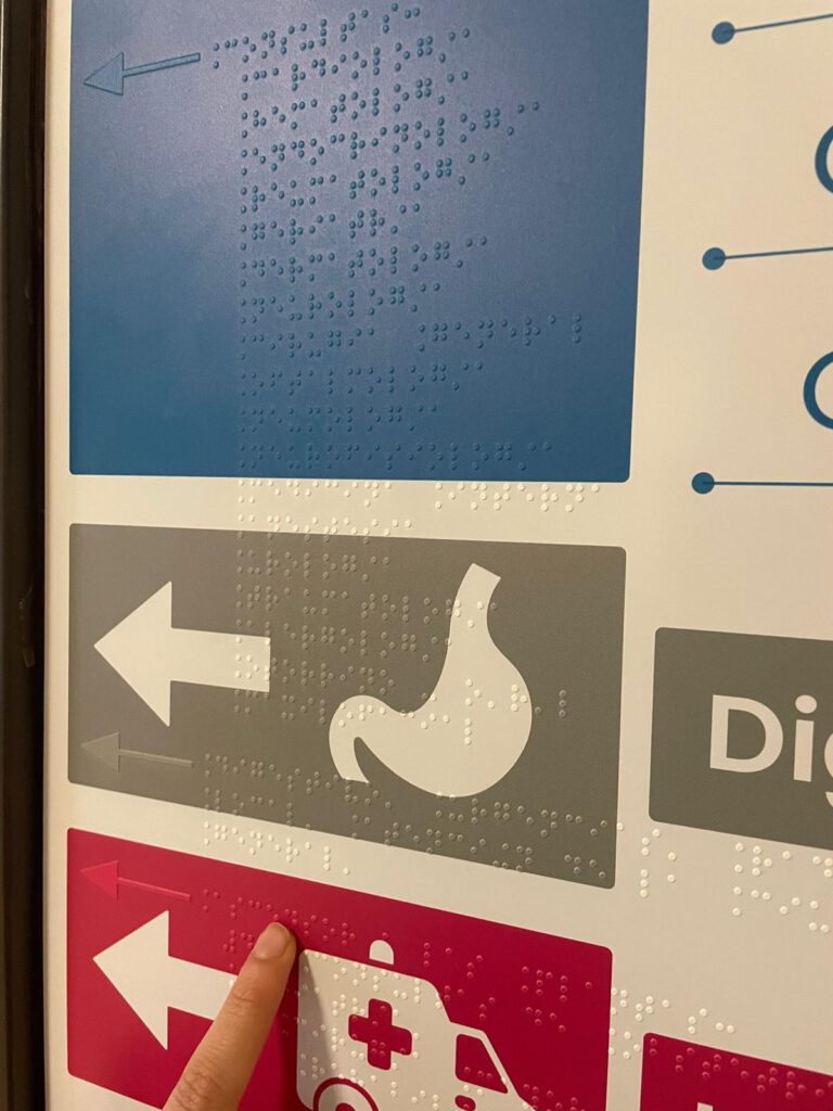 Primer plano de un panel de señalización accesible con flechas direccionales e iconos en relieve y braille. Una mano señala con el dedo una línea de braille sobre una franja roja; encima se ven otras franjas azul y gris con flechas hacia la izquierda.