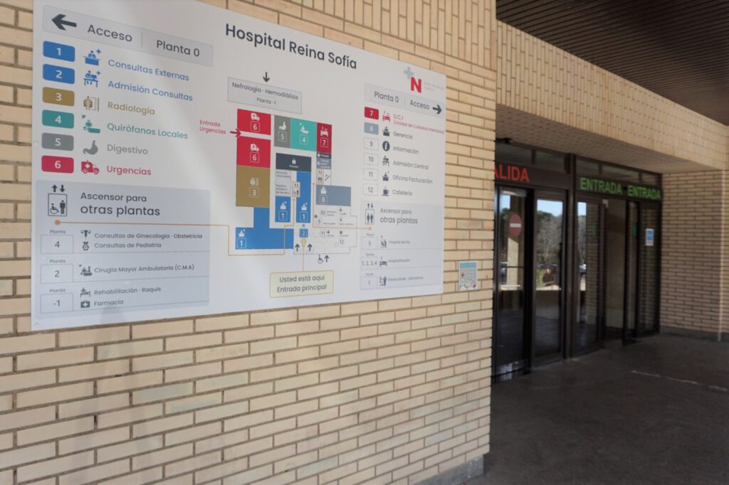 Panel grande de señalización del Hospital Reina Sofía en una pared de ladrillo, con plano y listado de servicios por plantas (Consultas externas, Admisión, Radiología, Quirófanos locales, Digestivo y Urgencias), además de pictogramas de accesibilidad y la indicación “Usted está aquí”. A la derecha se ve la entrada acristalada del hospital con los rótulos “Entrada” y “Salida”.