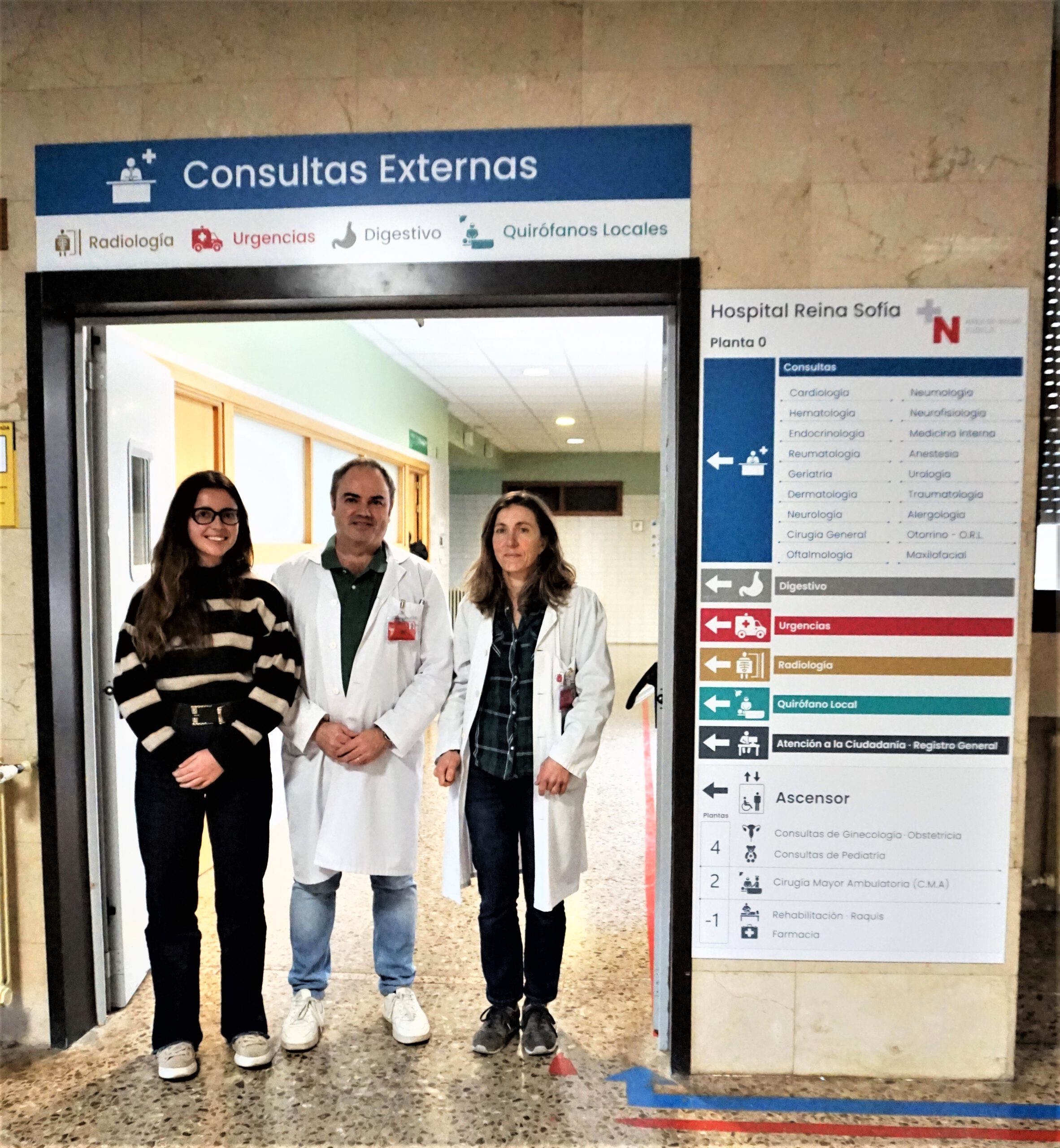 Entrada a la zona de “Consultas Externas” de un hospital: tres personas posan en la puerta, bajo un cartel azul con el rótulo principal y especialidades (Radiología, Urgencias, Digestivo, Quirófanos locales). A la derecha hay un directorio del Hospital Reina Sofía con la lista de servicios y señalización por colores.