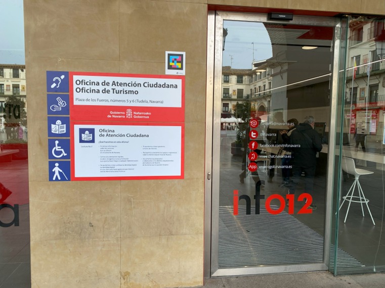 Panel de señalización accesible de la Oficina de Atención Ciudadana y Oficina de Turismo, junto a la puerta de entrada de vidrio con el texto “info112”.
