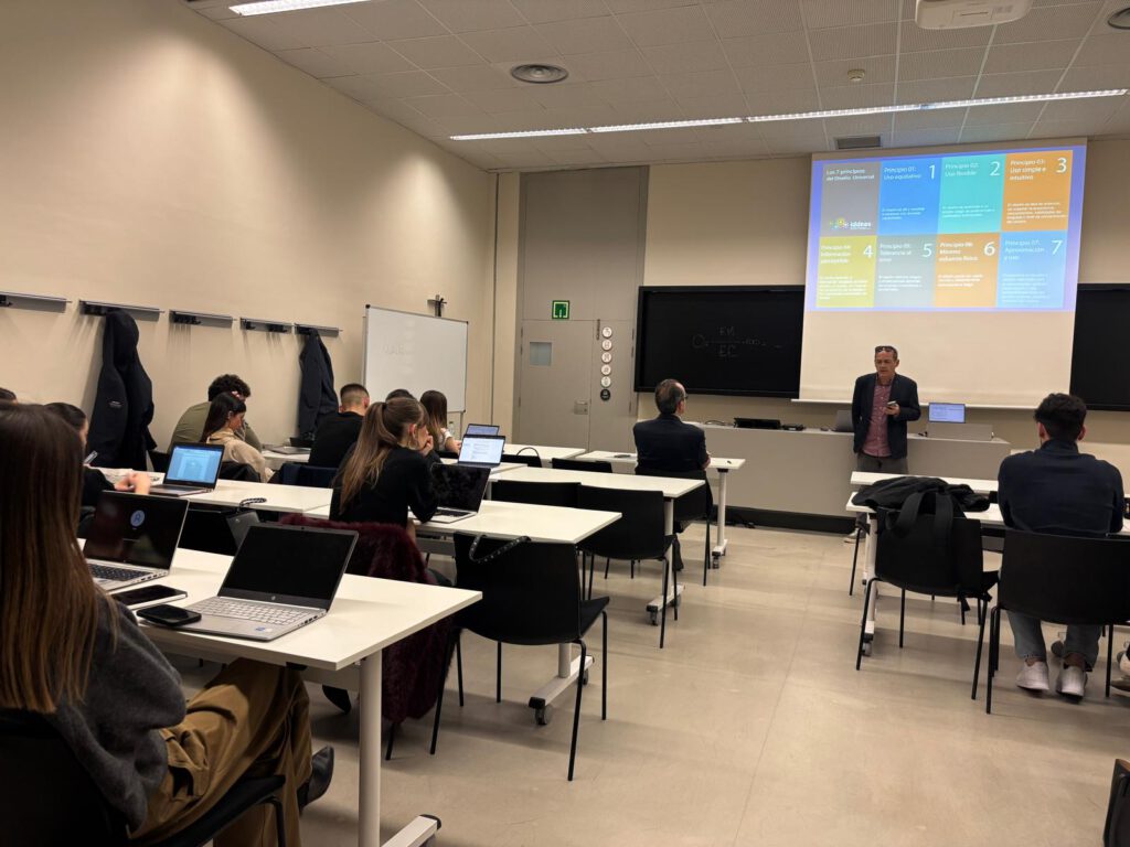 Clase de Marketing inclusivo con varias personas sentadas en mesas con ordenadores portátiles, asistiendo a una formación. Al frente, una persona expone mientras se proyecta una diapositiva sobre los principios del diseño universal. El público está distribuido en filas, prestando atención o tomando notas. El espacio cuenta con pizarra, pantalla de proyección y percheros en la pared.