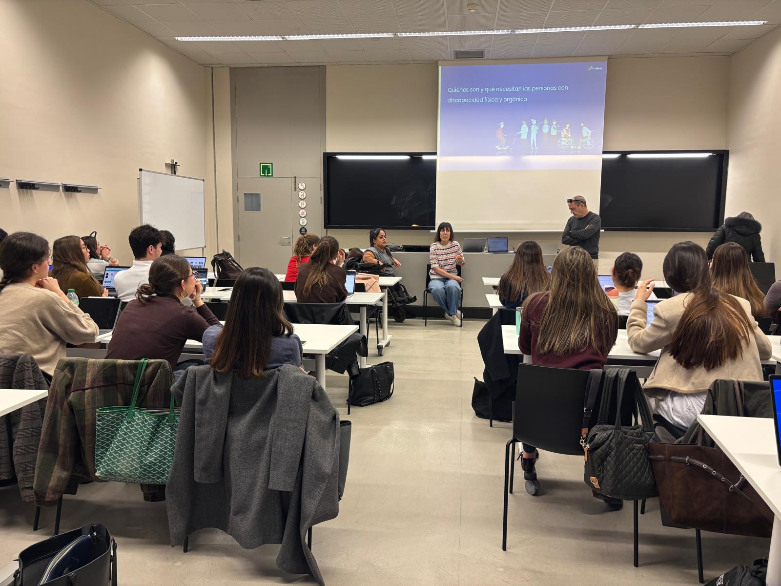 Clase de Marketing inclusivo con varias personas sentadas en mesas, asistiendo a una formación. Al fondo, dos personas intervienen frente al grupo mientras se proyecta una presentación sobre discapacidad física y orgánica. El público presta atención, algunas personas toman notas o utilizan ordenadores portátiles. El espacio es amplio, con pizarra y pantalla de proyección.